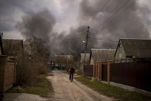 A Ukrainian man rides his bicycle near to a factory and a store burning after been bombarded in Irpin, on the outskirts of Kyiv, Ukraine, Sunday, March 6, 2022. (AP Photo/Emilio Morenatti)