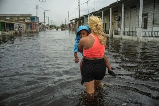 A woman carries a child as she wades through a street flooded in the passing of Hurricane Helene, in Batabano, Mayabeque province, Cuba, Sept. 26, 2024. (AP Photo/Ramon Espinosa, File)