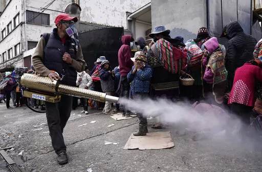 A worker sprays disinfectant around people who traveled from the provinces to Monte de Oración church for donated food and clothing during the church's annual Good Samaritan campaign in La Paz, Bolivia, Tuesday, Dec. 28, 2021. Due to the COVID-19 pandemic, the church is distributing the goods outdoors. (AP Photo/Freddy Barragan)