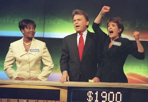 Johnna Goodwin, left, watches as Donna Handel, right, reacts after winning on "Wheel of Fortune," with host Pat Sajak during a taping of the game show in Philadelphia on April 17, 1999. The game show was in Philadelphia for the weekend taping a week's worth of shows. Ryan Seacrest will replace the retiring Sajak as host of “Wheel of Fortune.” (AP Photo/William Thomas Cain, File)