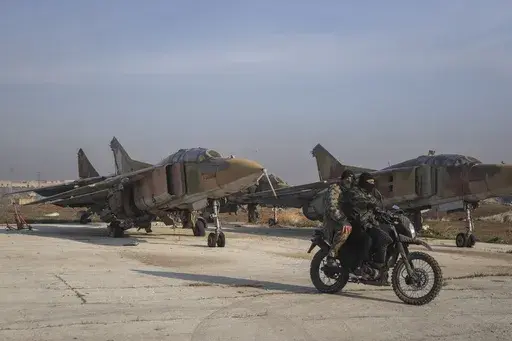 Syrian opposition fighters ride on a motorcycle past Syrian army planes at the Al-Nayrab military airport after they took control of the facility in the outskirts of Aleppo, Syria, Monday, Dec. 2, 2024. (AP Photo/Omar Albam)