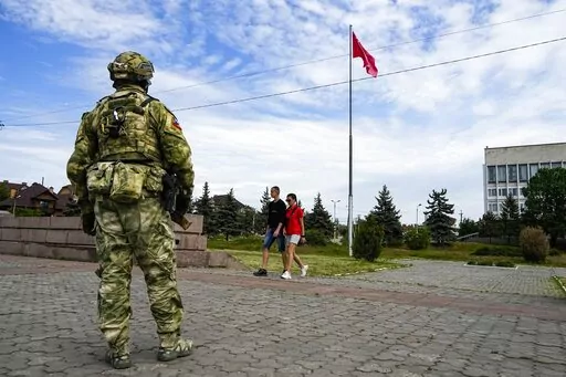 A young couple walks past a Russian soldier guarding an area at the Alley of Glory exploits of the heroes - natives of the Kherson region, who took part in the liberation of the region from the Nazi invaders, in Kherson, Kherson region, south Ukraine, Friday, May 20, 2022, with a replica of the Victory banner marking the 77th anniversary of the end of World War II right in the background. Ukrainian forces pressing an offensive in the south have zeroed in on Kherson, a provincial capital that has