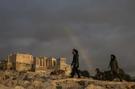 In this Wednesday, March 31, 2021, file photo, women wearing face masks walk with their dogs at Areopagus hill, in front of ancient Acropolis hill, as a rainbow is seen in the cloudy Athenian sky. Greece's culture ministry announced on Thursday, April 27, 2023, that pets will soon be allowed into more than 120 archaeological sites across the country, although not in some of the top tourist draws such as the Acropolis in Athens. (AP Photo/Petros Giannakouris, File)