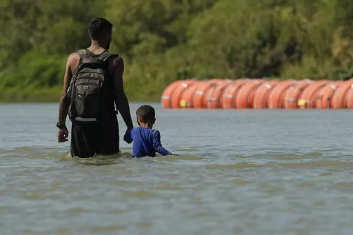 Migrants walk past large buoys being used as a floating border barrier on the Rio Grande, Aug. 1, 2023, in Eagle Pass, Texas. Texas attorneys asked federal appeals court judges Thursday, Oct. 5, 2023 to let the state keep in place a floating barrier of large concrete-anchored buoys to block migrants from crossing the Rio Grande — a barrier the Biden administration says was illegally deployed without needed federal authorization. (AP Photo/Eric Gay, file)